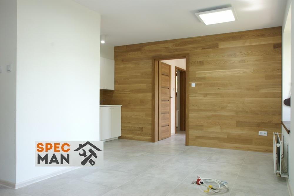 Wnętrze pokoju z drewnianą okładziną ścienną, szarą podłogą z płytek i częściowo widoczną zabudową kuchenną z białymi szafkami; widoczne gniazdka elektryczne i przewody na podłodze.