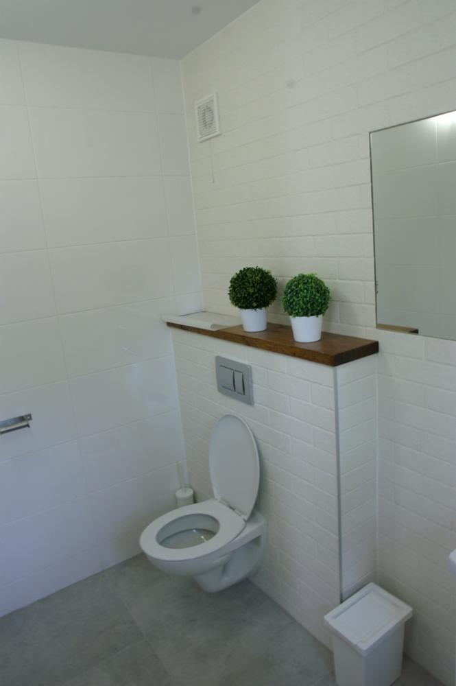 Nowoczesna łazienka z białymi płytkami imitującymi cegłę, podwieszana toaleta z ukrytą spłuczką, drewniana półka z dwoma małymi sztucznymi roślinami, szara podłoga z płytek.