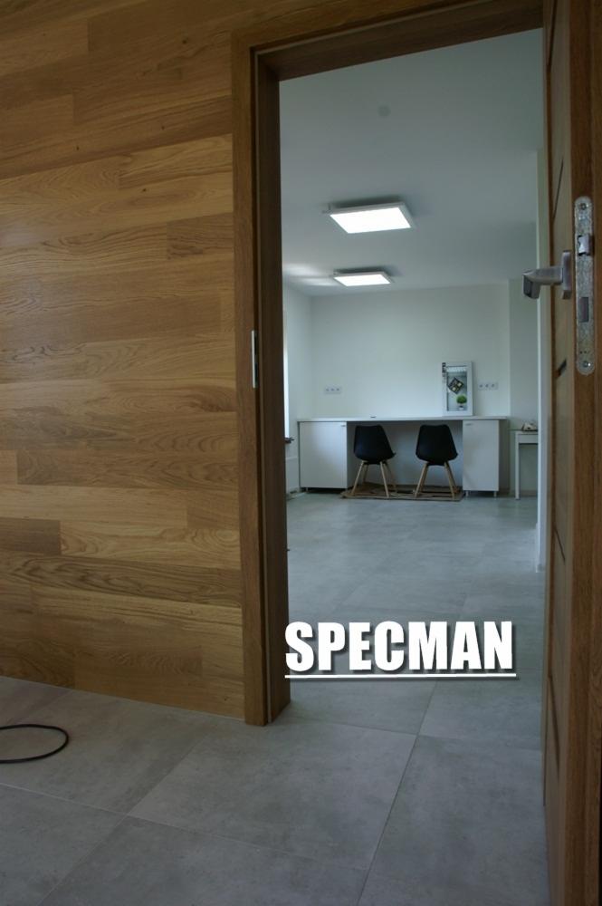 Widok przez otwarte drewniane drzwi na minimalistyczne biuro z drewnianą ścianą po lewej, szare płytki podłogowe, białe ściany, dwa czarne krzesła i białe biurko pod dwoma kwadratowymi lampami...