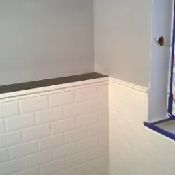 Fragment ściany łazienki z białymi, prostokątnymi płytkami ułożonymi w cegiełkę, szara ściana powyżej, ciemny blat i biała listwa dekoracyjna, narożnik z widocznym gniazdkiem elektrycznym,...