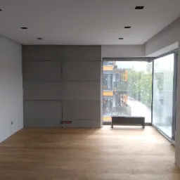 Wnętrze pokoju z ułożonym parkietem z jasnego drewna, szara ściana z płyt, duże okno z widokiem na zewnątrz i czarne punkty świetlne na suficie.