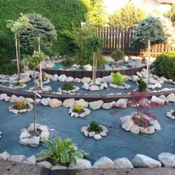 Nowo założony ogród z różnorodnymi roślinami, otoczonymi kamieniami i oddzielonymi drewnianymi belkami, na zielonej macie szkółkarskiej, z widocznym ogrodzeniem w tle.