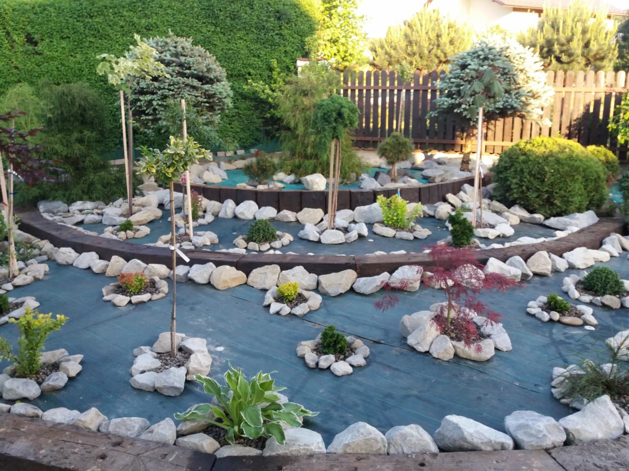 Nowo założony ogród z różnorodnymi roślinami, otoczonymi kamieniami i oddzielonymi drewnianymi belkami, na zielonej macie szkółkarskiej, z widocznym ogrodzeniem w tle.