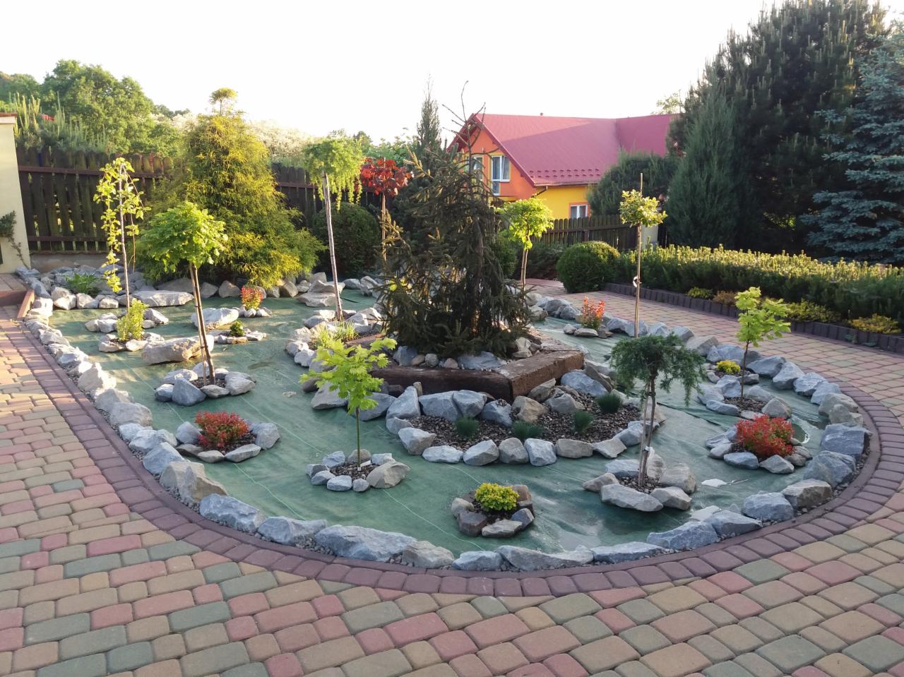 Ogród w trakcie aranżacji: rozłożona agrowłóknina, nasadzenia drzewek i krzewów otoczone kamieniami, obrzeże z kostki brukowej, w tle żółty dom z czerwonym dachem.