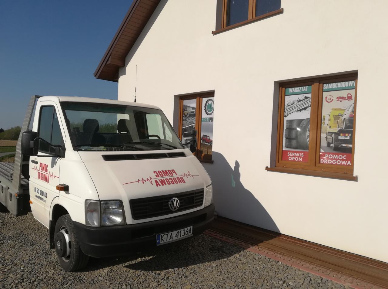 Biała laweta pomocy drogowej z czerwonym napisem zaparkowana przed budynkiem warsztatu samochodowego z naklejkami reklamowymi na oknach.