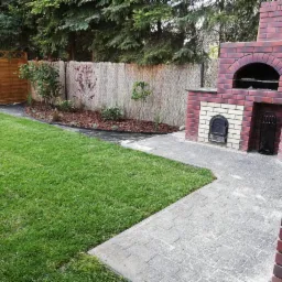 Zadbany ogród z równo przystrzyżonym trawnikiem, nowo posadzone rośliny na rabacie z korą, ścieżka z kostki brukowej prowadząca do murowanego grilla z cegły.