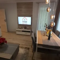 Salon z drewnianą podłogą, sofą z kwiatem, stołem jadalnym i telewizorem z logo firmy MAXBUD na ścianie z drewnopodobną okładziną.