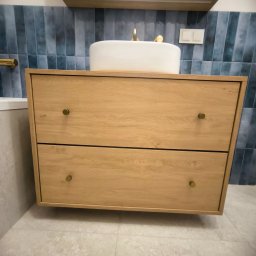 ERG INVESTMENTS Sp. z o.o. - Drewniana szafka pod umywalkę z dwoma szufladami i okrągłą umywalką nablatową. W tle niebieskie, pionowe płytki i lustro z oświetleniem.