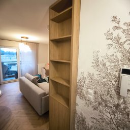 ERG INVESTMENTS Sp. z o.o. - Drewniany regał z półkami i szafką, wkomponowany w ścianę z motywem drzewa. Widok na salon z sofą i balkonem. Naturalne światło.