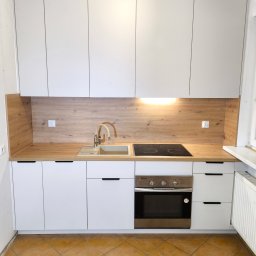 ERG INVESTMENTS Sp. z o.o. - Nowoczesna kuchnia z białymi szafkami i drewnianym blatem. Widoczny zlew, płyta indukcyjna i piekarnik. Minimalistyczny design z ciemnymi uchwytami.