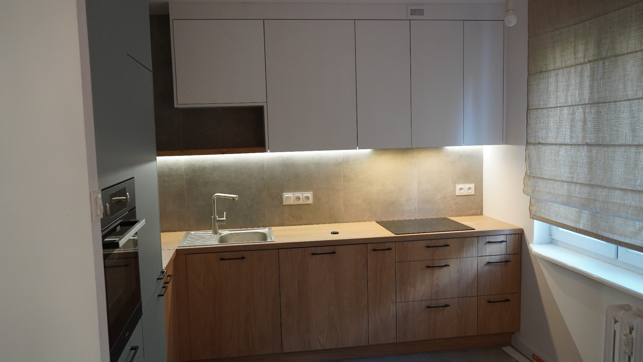 Nowoczesna kuchnia z drewnianymi szafkami dolnymi z czarnymi uchwytami, białymi szafkami górnymi, szarym blatem i oświetleniem LED pod szafkami, widok na zlew, płytę indukcyjną i roletę rzymską...