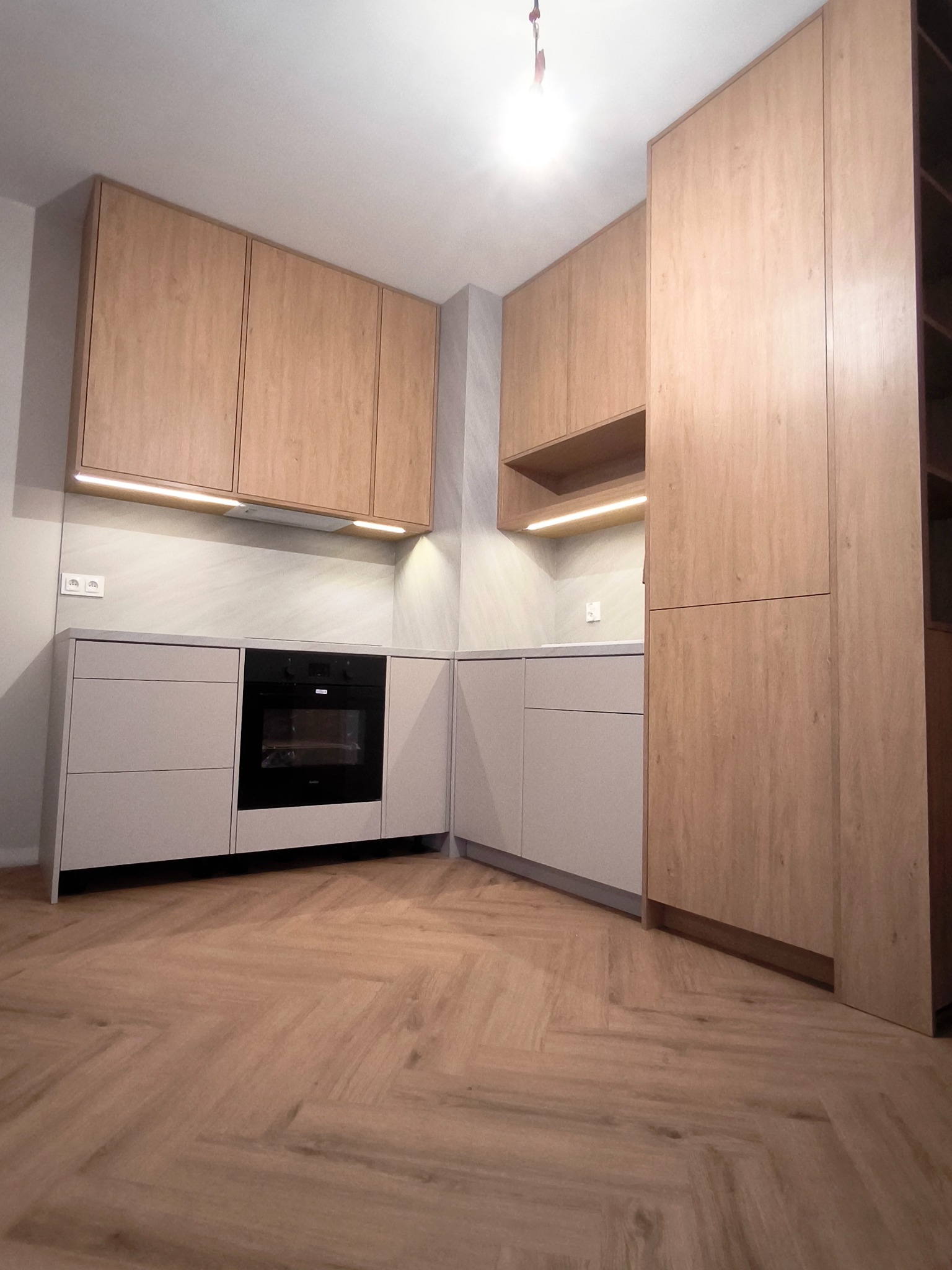 Jasna kuchnia z drewnianymi frontami i oświetleniem LED pod szafkami wiszącymi, widok na zabudowę kuchenną z piekarnikiem i podłogą w jodełkę.