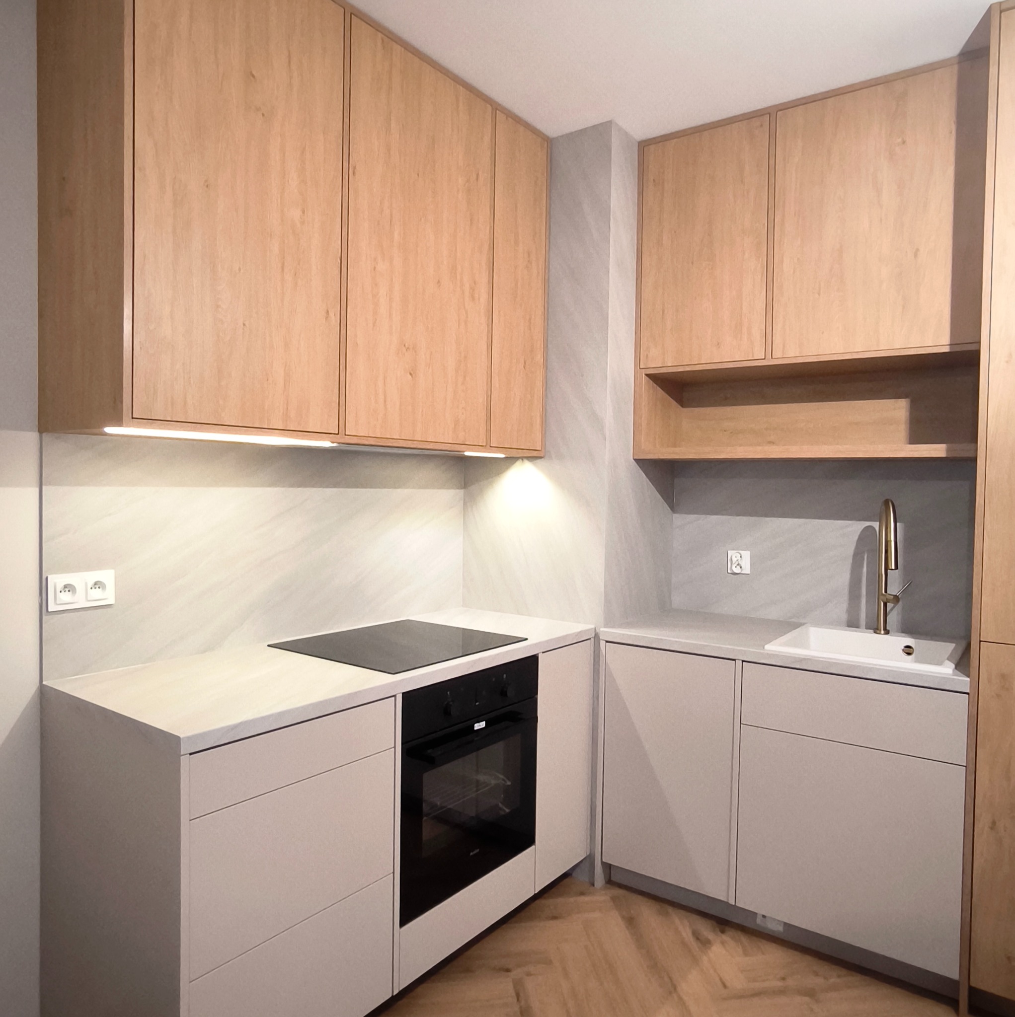 Jasna kuchnia na wymiar z drewnianymi szafkami górnymi i beżowymi dolnymi. Widoczna płyta indukcyjna, piekarnik i złota bateria przy zlewie. Parkiet w jodełkę na podłodze.