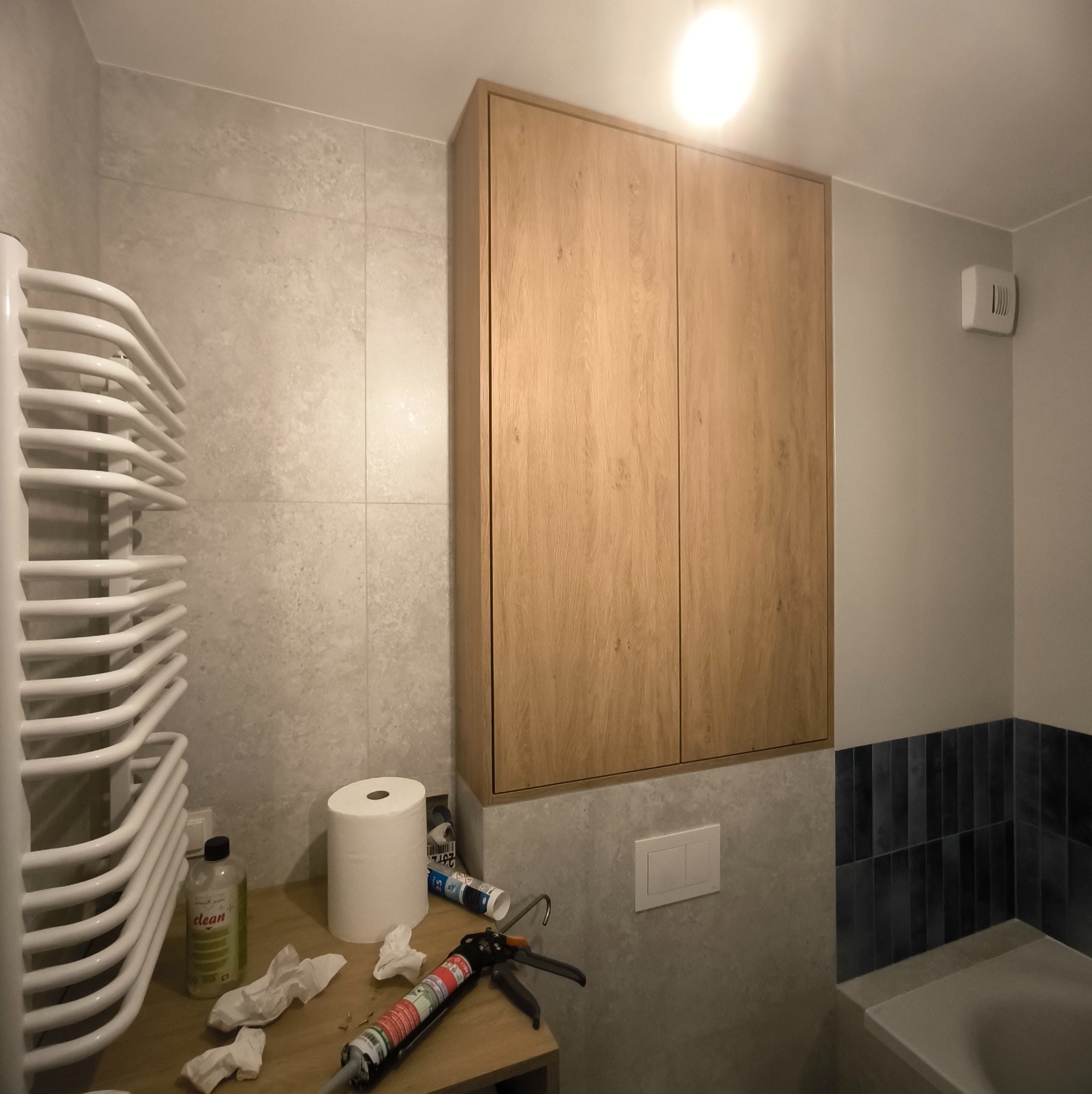 Drewniana szafka łazienkowa nad toaletą, z widocznym grzejnikiem, narzędziami i akcesoriami montażowymi. Ujęcie z bliska, prezentujące detale wykończenia.