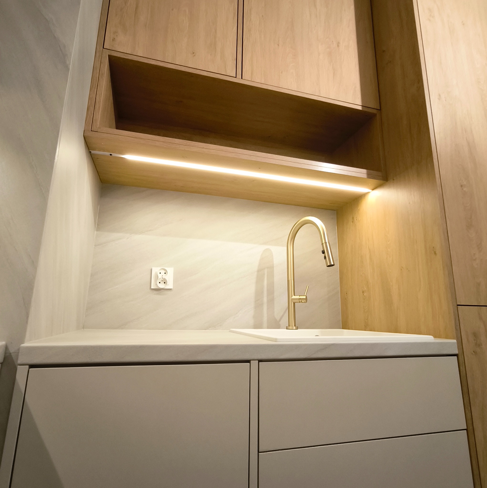 Nowoczesna kuchnia z drewnianymi szafkami, złotą baterią i podświetleniem LED. Minimalistyczny design, jasne kolory i gładkie powierzchnie. Widoczny fragment blatu i gniazdko elektryczne.