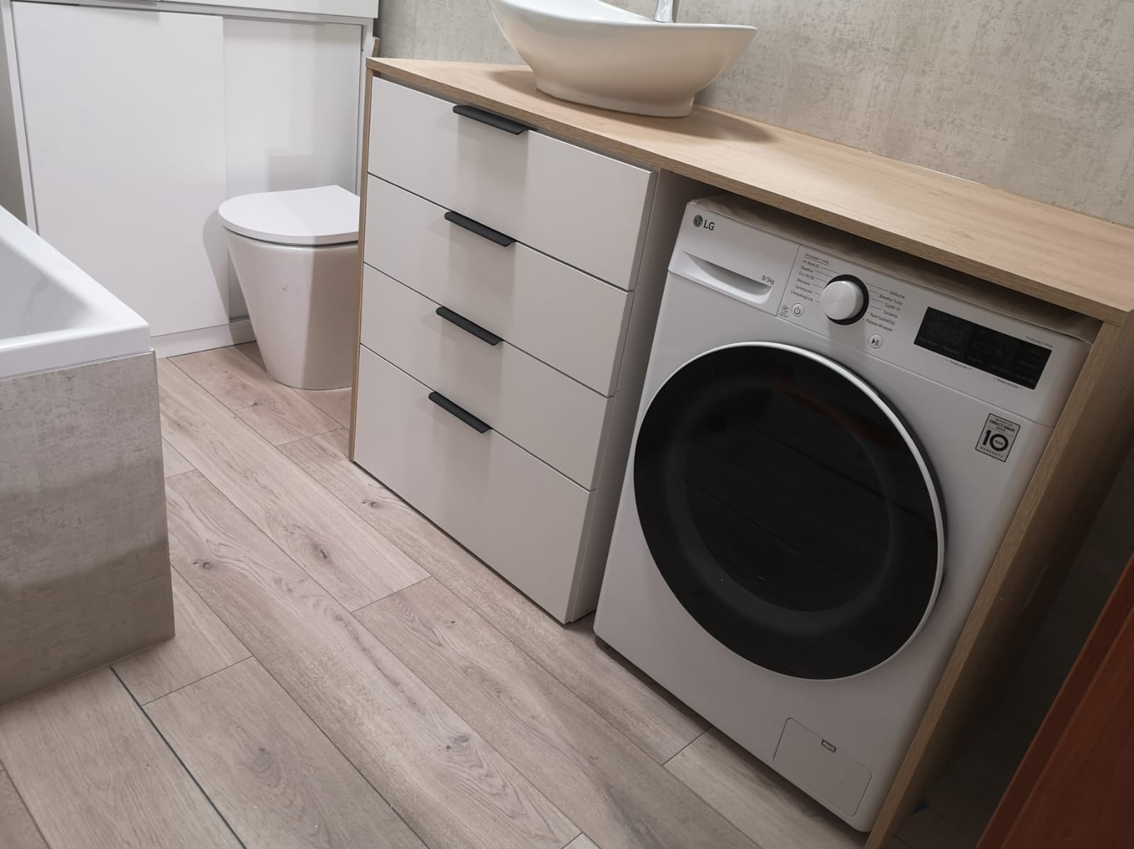 Nowoczesna łazienka z białą umywalką nablatową, szafką z szufladami i pralką LG w zabudowie z blatem drewnopodobnym. Jasne drewniane panele podłogowe.