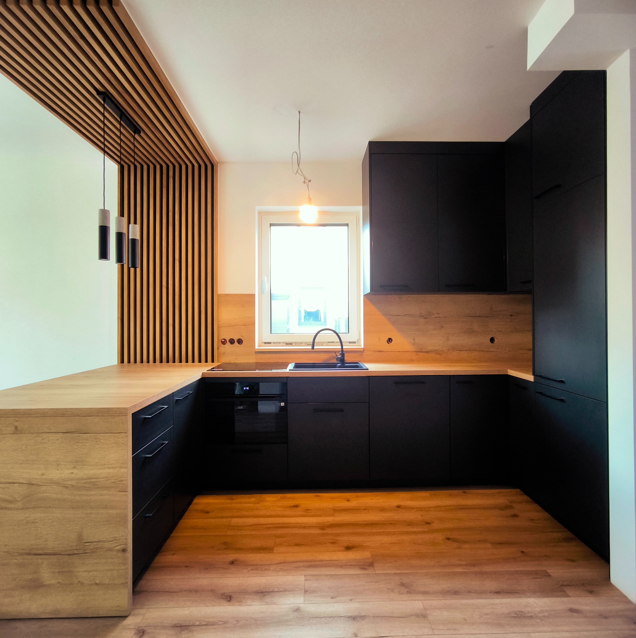 Nowoczesna kuchnia w Opalenicy z czarnymi szafkami i drewnianym blatem. Widoczne oświetlenie i drewniane lamele na ścianie. Minimalistyczny design.