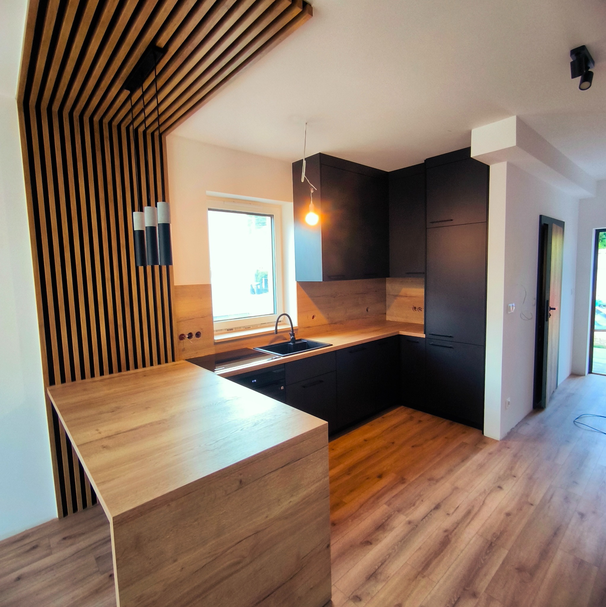 Nowoczesna kuchnia z czarnymi szafkami i drewnianym blatem, oświetlona wiszącymi lampami. Dekoracyjna ściana z drewnianych listew dodaje unikalnego charakteru.