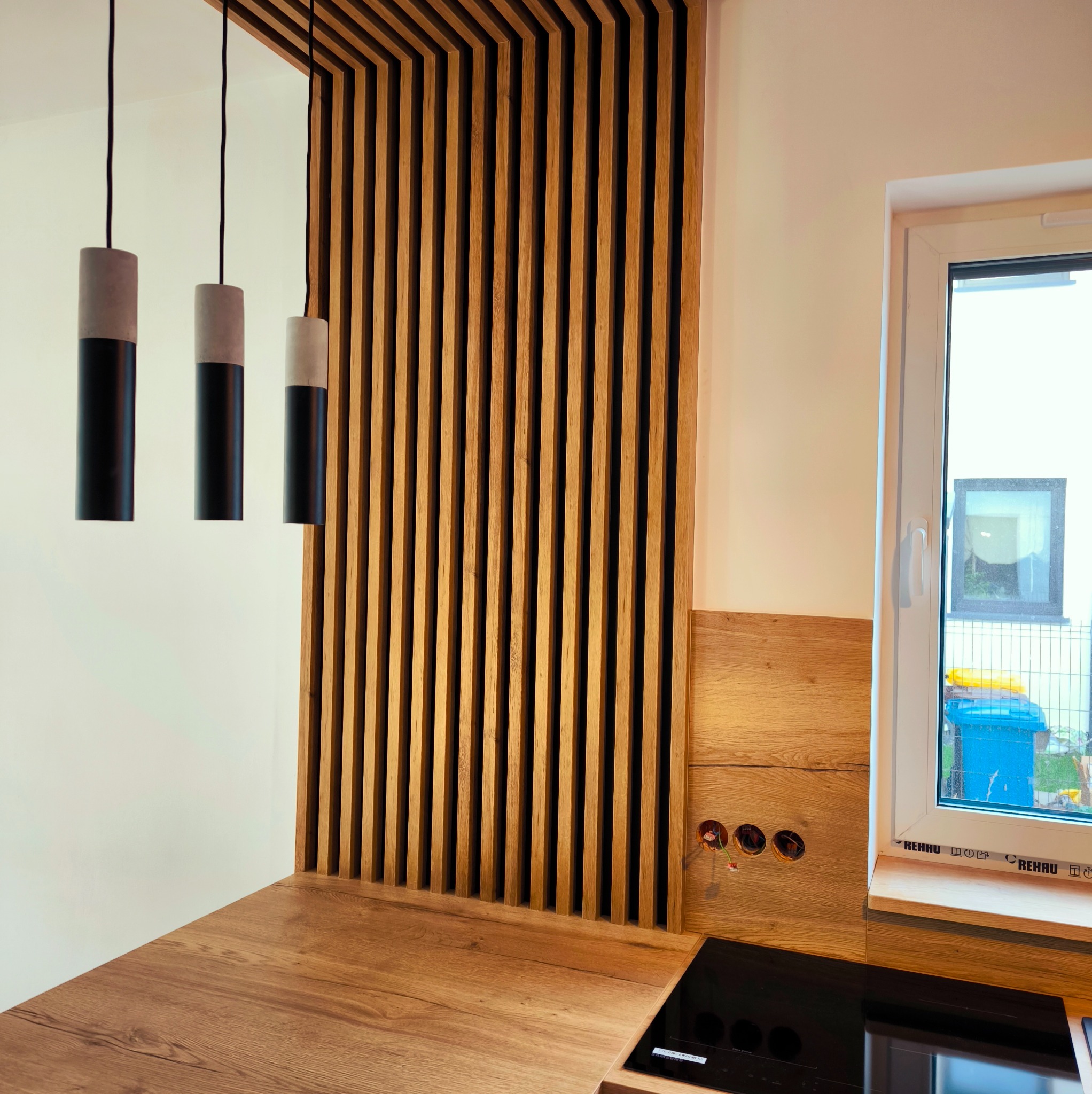 Nowoczesna kuchnia z drewnianym blatem, lamelami i lampami wiszącymi. Widoczne gniazdka elektryczne i okno z widokiem na zewnątrz. Minimalistyczny design.