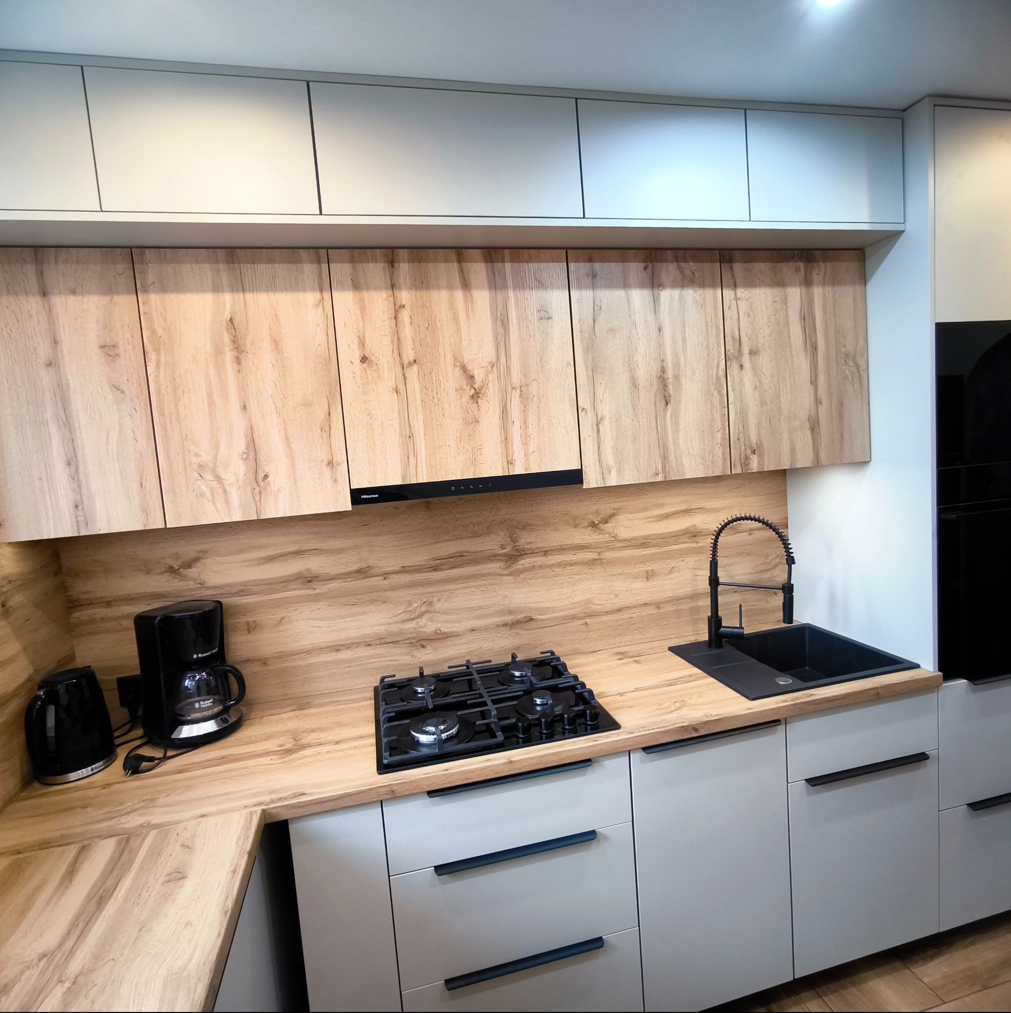 Nowoczesna kuchnia z drewnianym blatem i szarymi szafkami. Czarny zlew i bateria, płyta gazowa. Ekspres do kawy i czajnik na blacie. Górne szafki białe i drewniane.