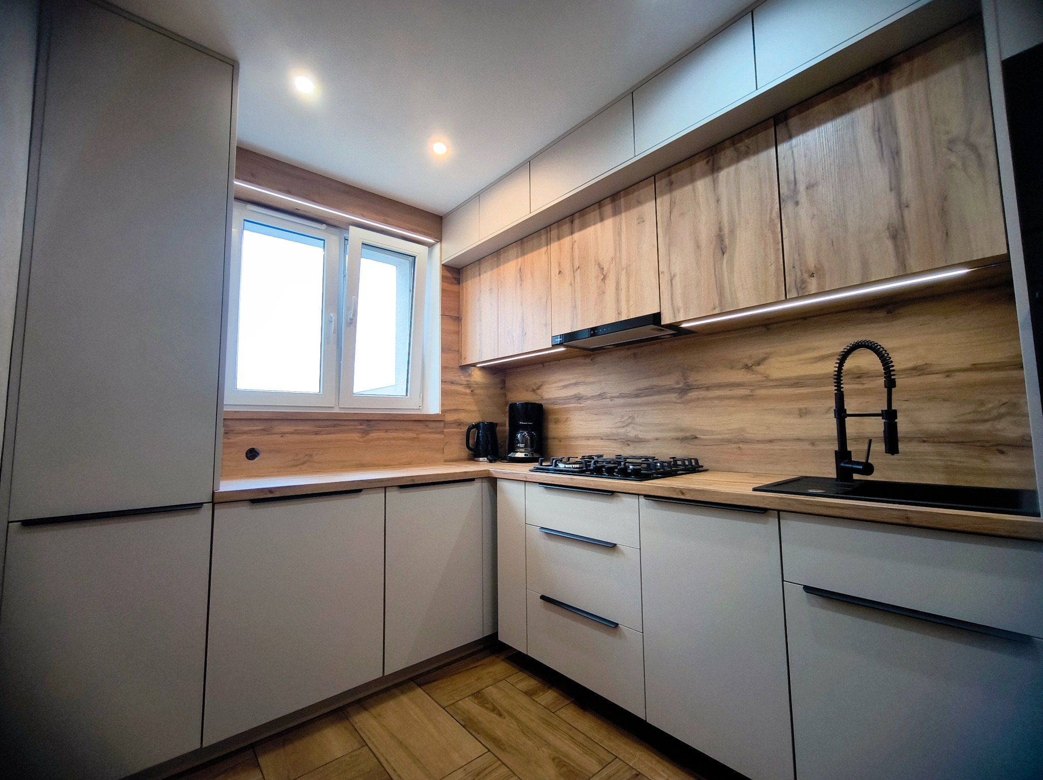 Nowoczesna kuchnia z drewnianym blatem i frontami w odcieniach szarości. Widoczna płyta gazowa, czarny kran i oświetlenie LED pod szafkami.