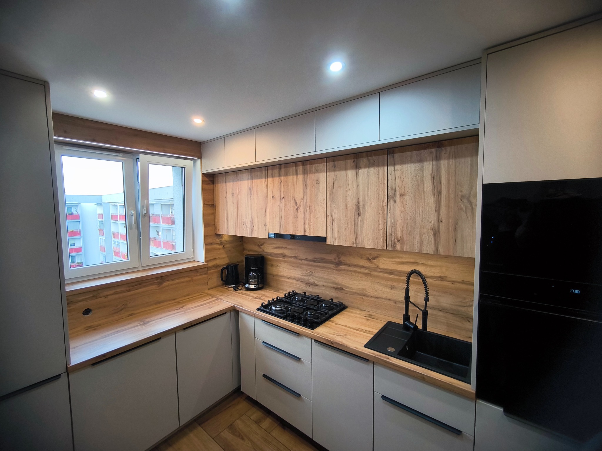 Nowoczesna kuchnia z drewnianym blatem i szarymi szafkami. Widoczny zlew, płyta gazowa i okno z widokiem na budynek. Minimalistyczny design, ciepłe oświetlenie.