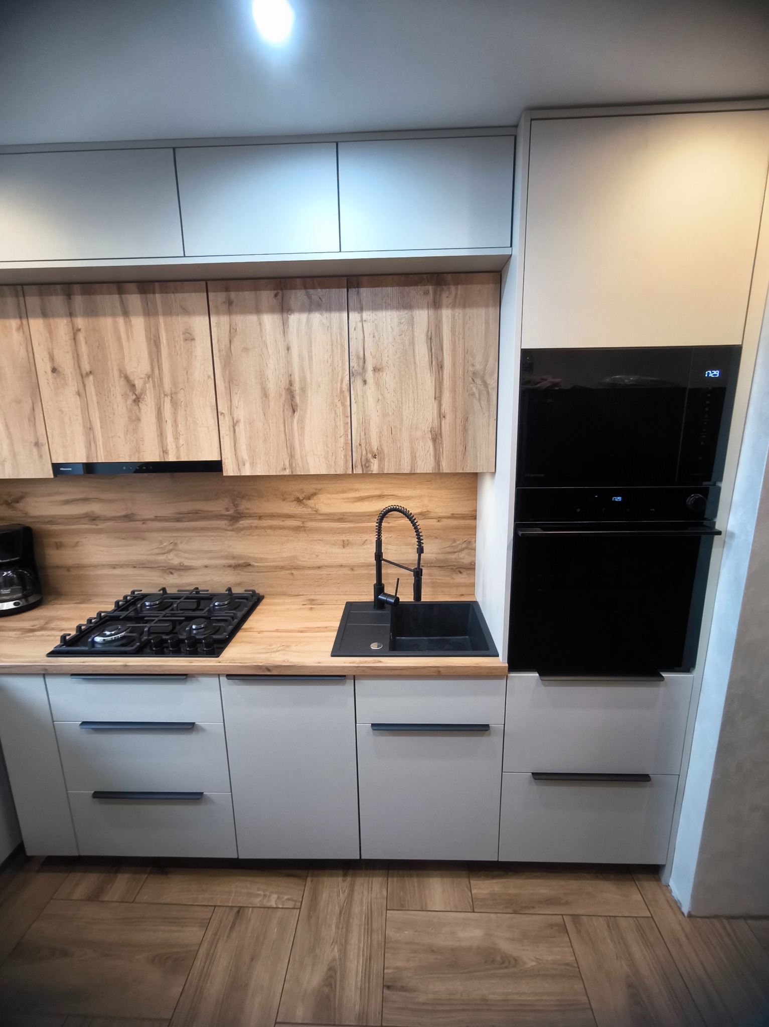 Nowoczesna kuchnia z drewnianymi akcentami: jasne szafki, drewnopodobny blat i górne szafki, czarny zlew i piekarnik. Minimalistyczny design, ciepłe oświetlenie.