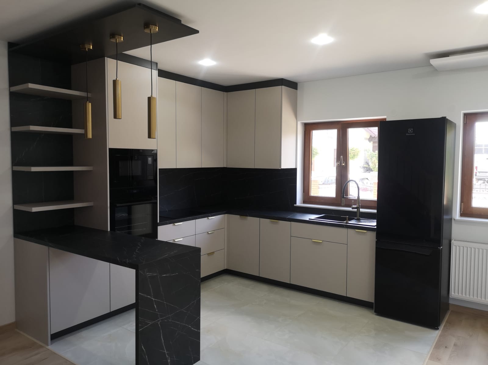 Nowoczesna kuchnia z beżowymi szafkami, czarnym blatem imitującym marmur i złotymi lampami wiszącymi. Widoczne okno i czarna lodówka Electrolux.