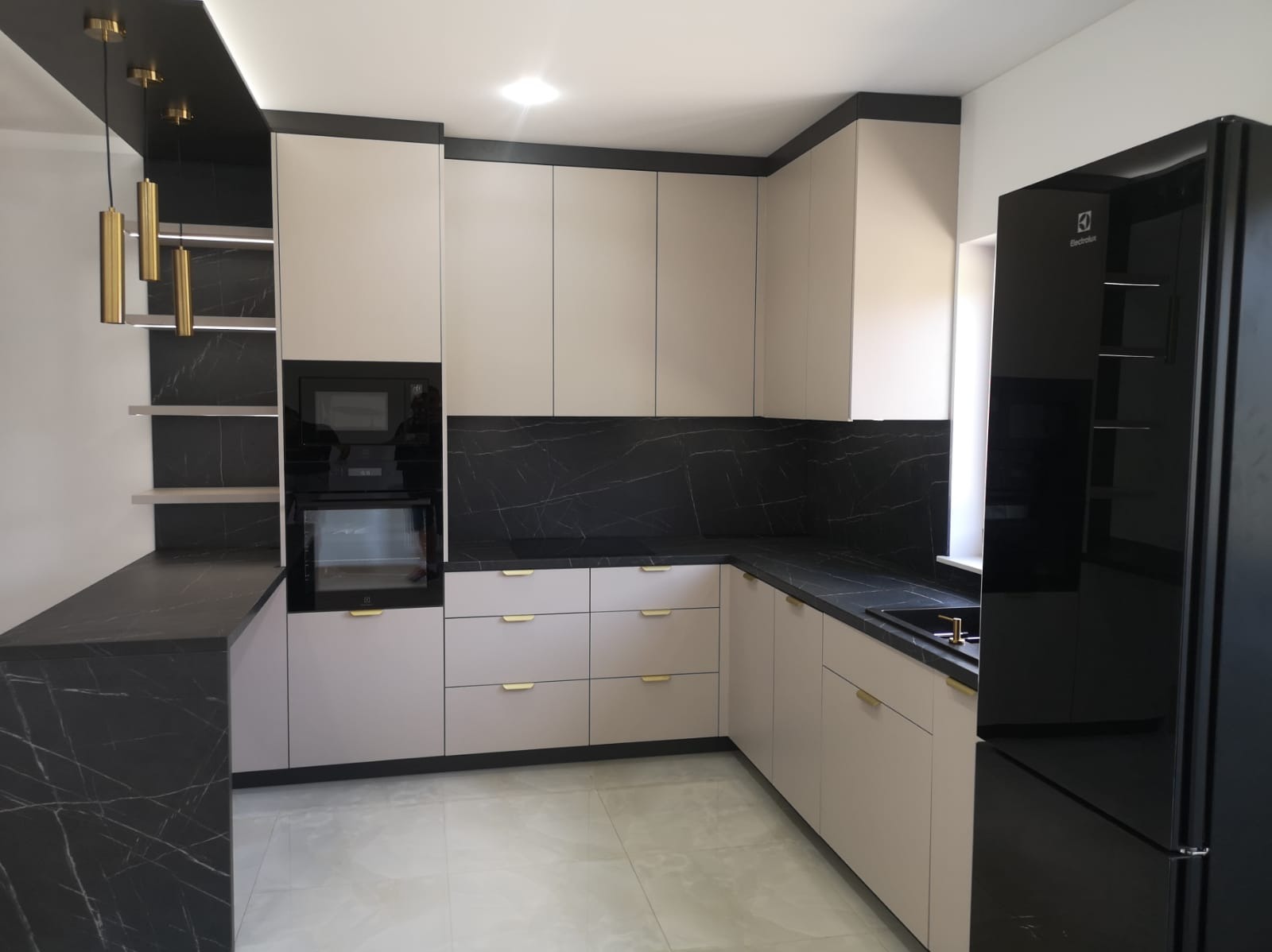Nowoczesna kuchnia z beżowymi szafkami, czarnym blatem imitującym marmur i złotymi uchwytami. Czarna lodówka Electrolux. Oświetlenie punktowe i wiszące lampy.