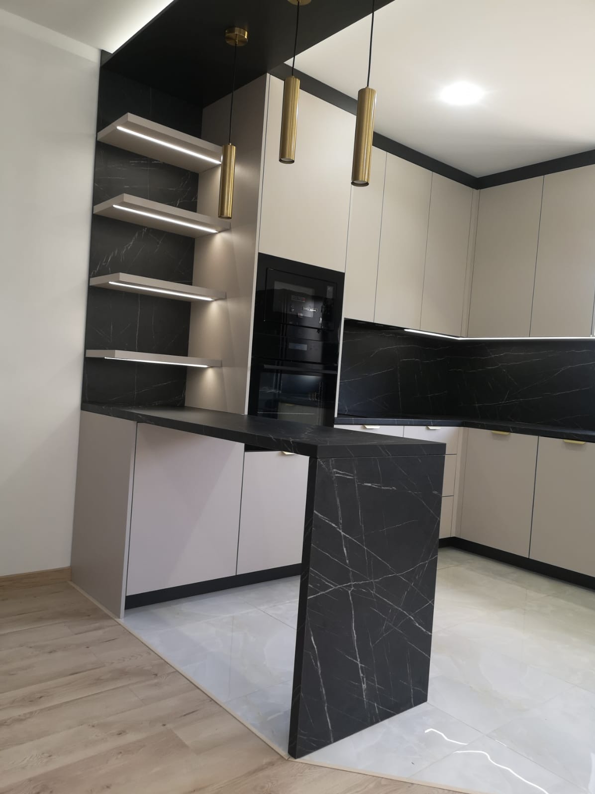 Nowoczesna kuchnia na wymiar z czarnym blatem imitującym marmur i podświetlanymi półkami. Jasne fronty szafek i złote lampy wiszące dodają elegancji.