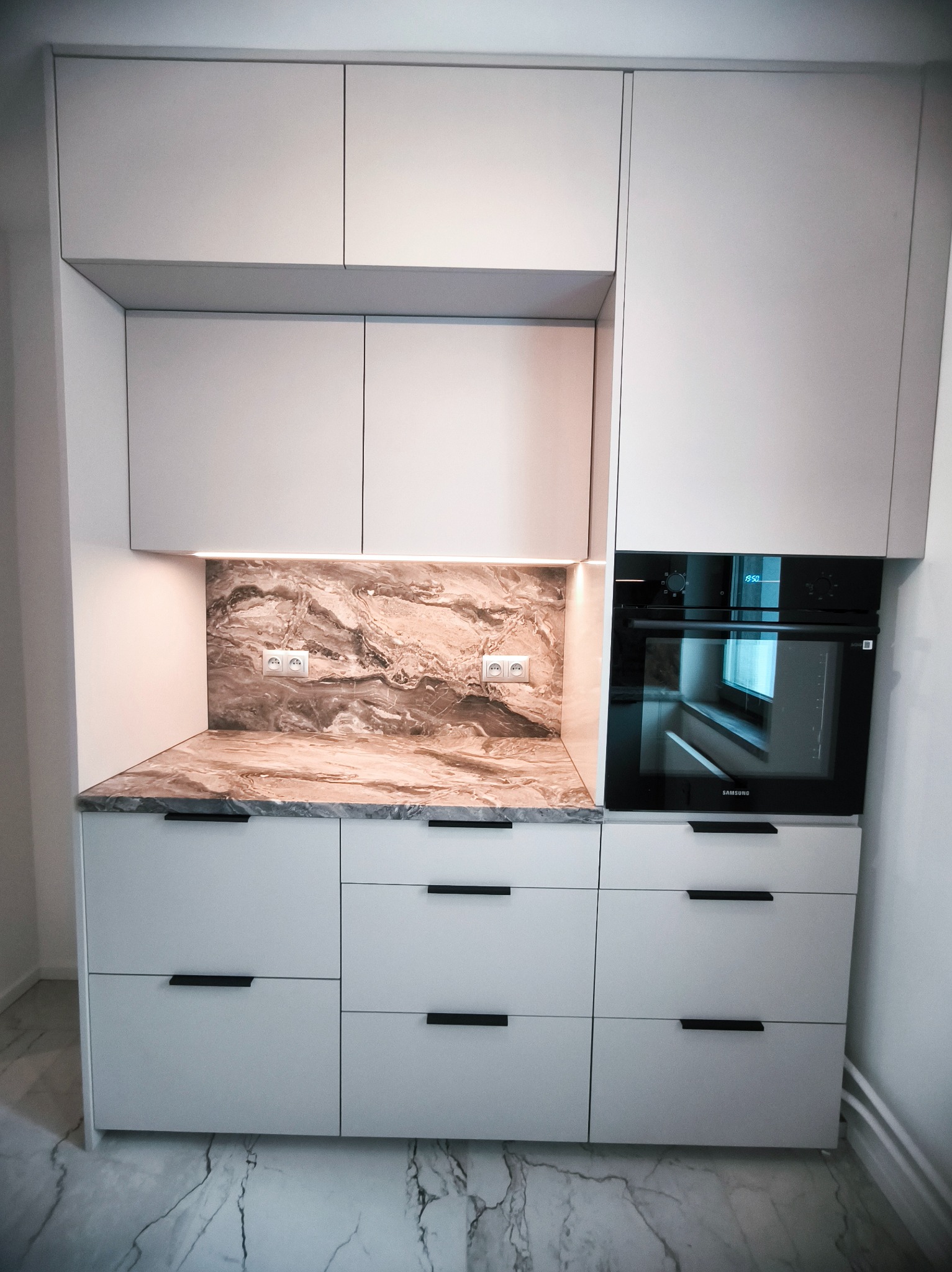 Jasna zabudowa kuchenna z marmurowym blatem i piekarnikiem Samsung. Minimalistyczne uchwyty, oświetlenie LED podszafkowe, marmurowa podłoga.