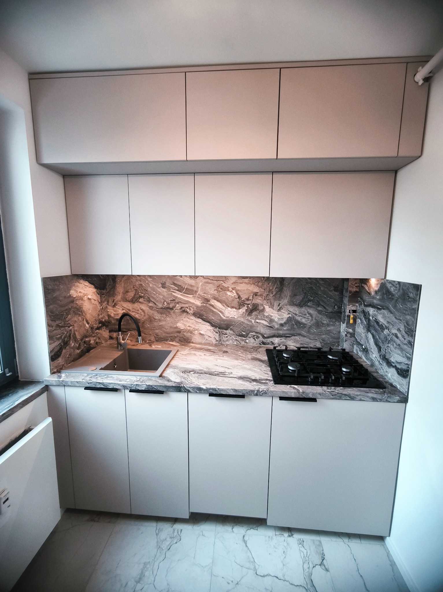 Jasna kuchnia z marmurowym blatem i czarną płytą gazową. Minimalistyczny design, gładkie fronty szafek, nowoczesne wykończenie. Ujęcie z dołu, widoczna marmurowa podłoga.