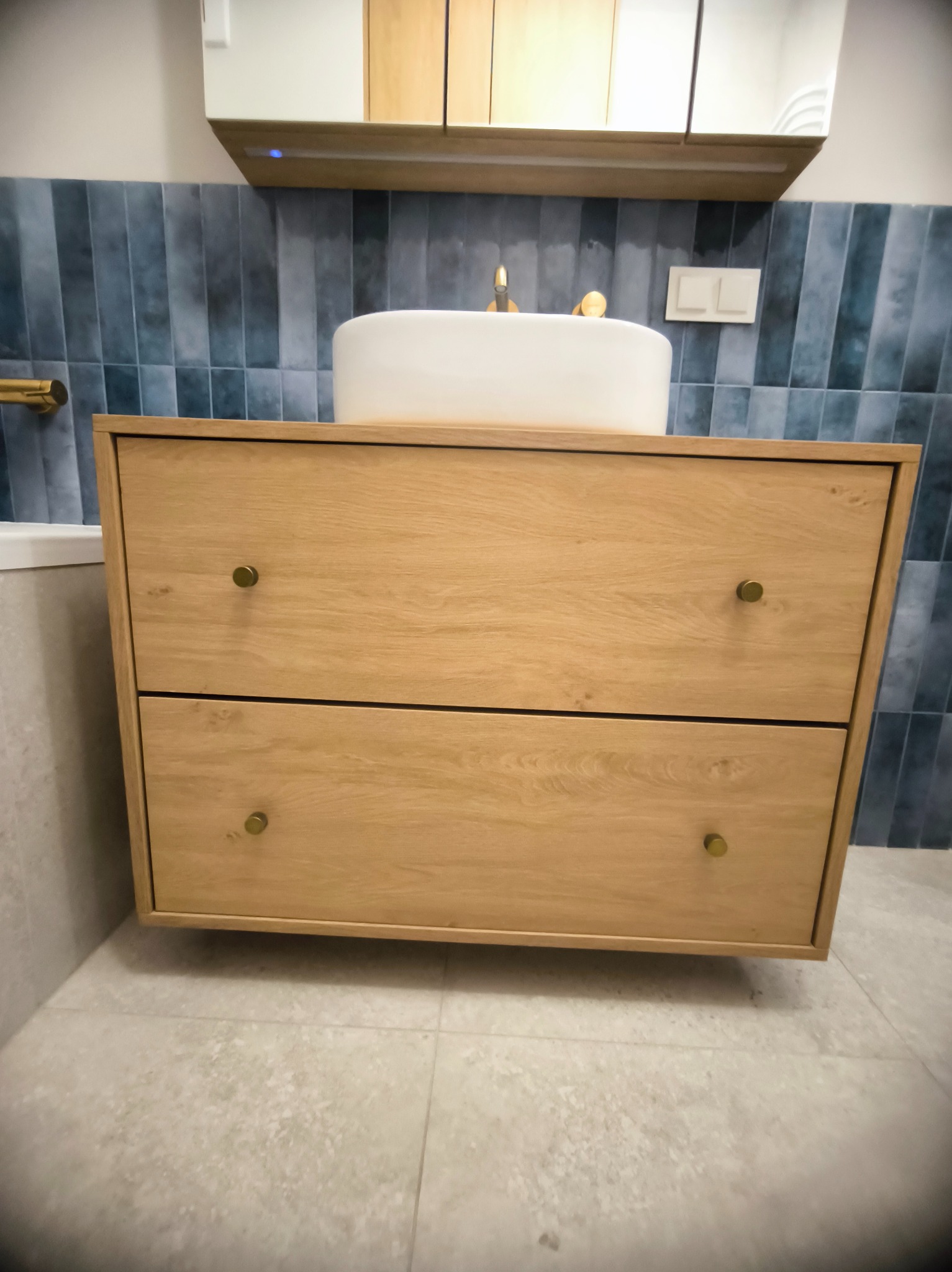 Drewniana szafka pod umywalkę z dwoma szufladami i okrągłą umywalką nablatową. W tle niebieskie, pionowe płytki i lustro z oświetleniem.