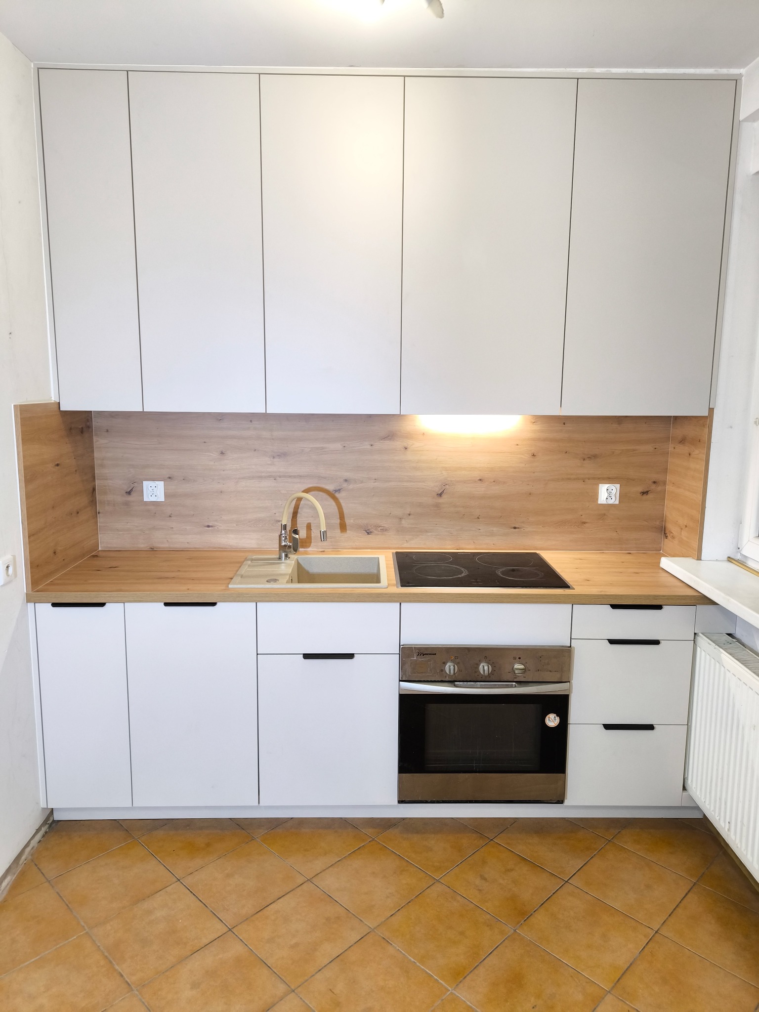 Nowoczesna kuchnia z białymi szafkami i drewnianym blatem. Widoczny zlew, płyta indukcyjna i piekarnik. Minimalistyczny design z ciemnymi uchwytami.