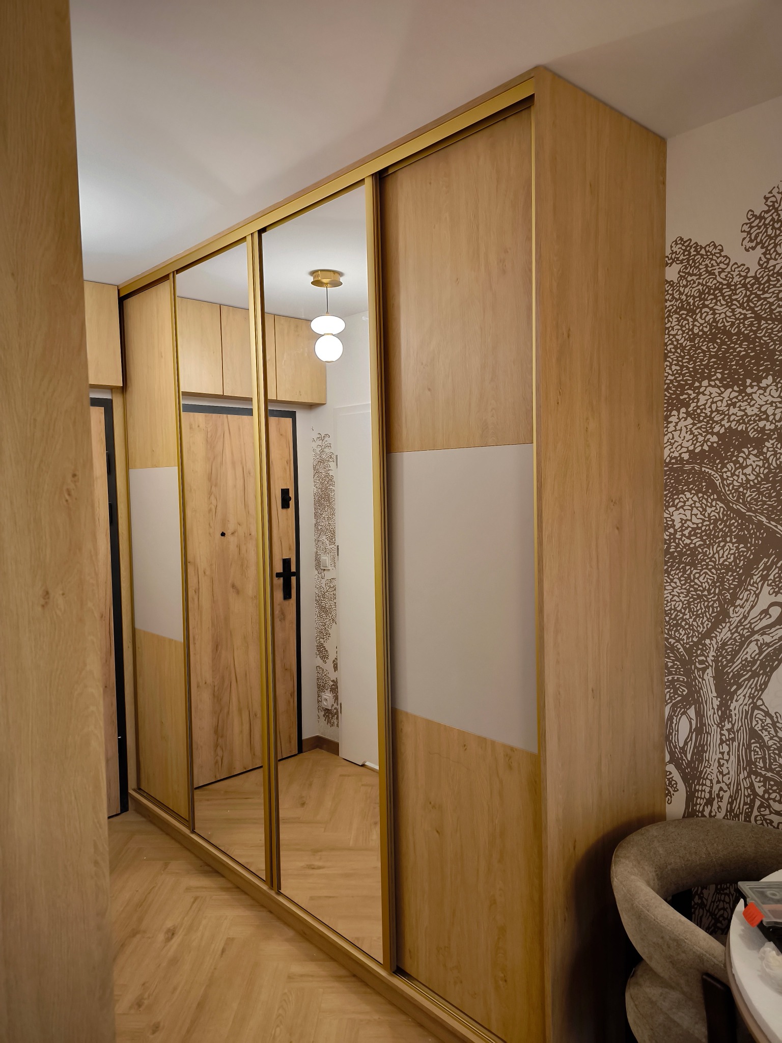 Jasna szafa wnękowa z przesuwnymi lustrzanymi drzwiami i złotymi ramami, dopasowana do drewnianej podłogi i drzwi wejściowych. Na ścianie dekoracyjny motyw drzewa.