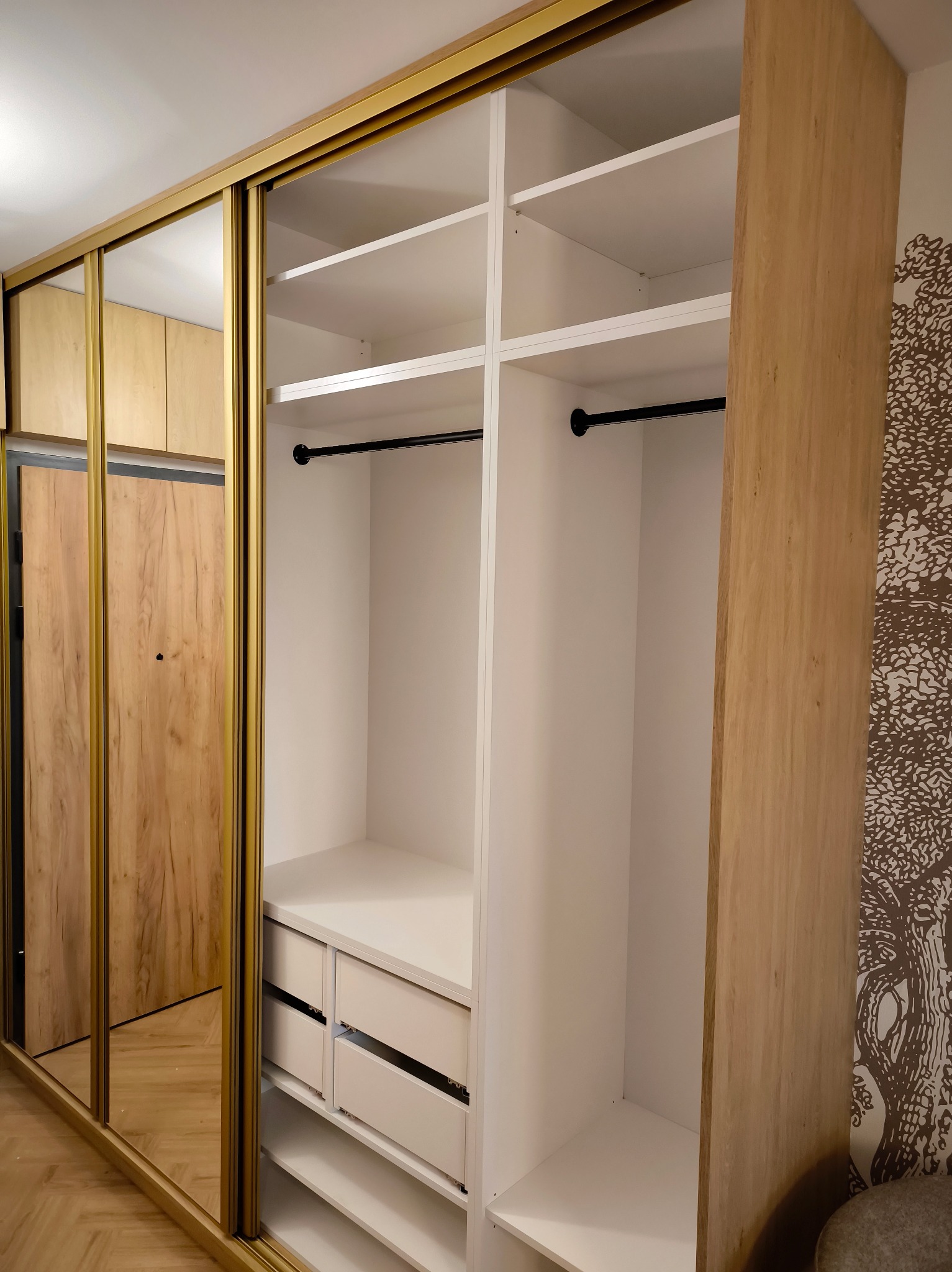 Szafa wnękowa z przesuwnymi drzwiami lustrzanymi w złotej ramie, wnętrze z półkami, szufladami i drążkiem na ubrania. Na ścianie tapeta z motywem drzewa.