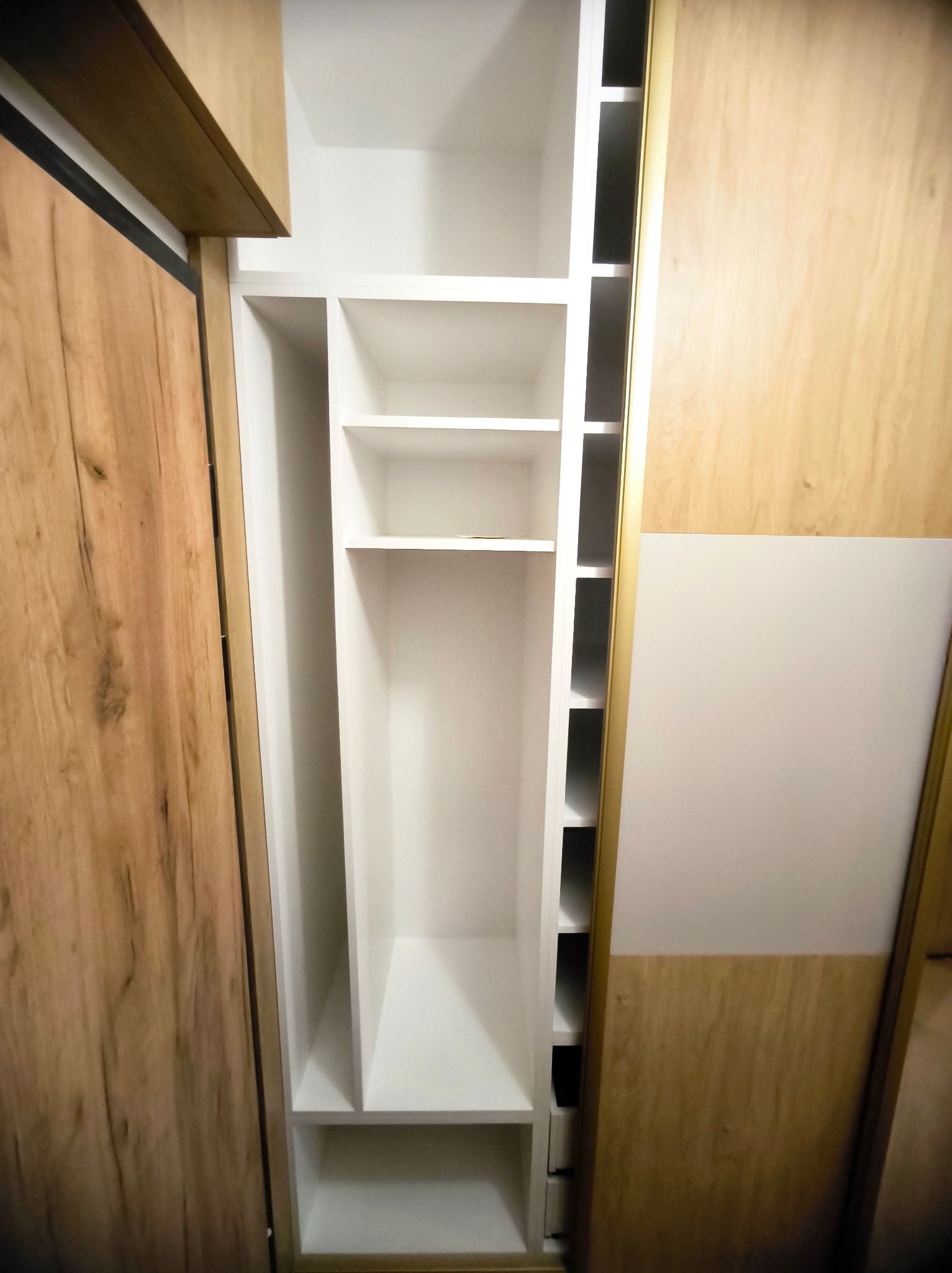 Wnętrze szafy wnękowej z przesuwnymi drzwiami w odcieniach bieli i drewna. Widoczne półki i przegródki, tworzące funkcjonalną przestrzeń do przechowywania.