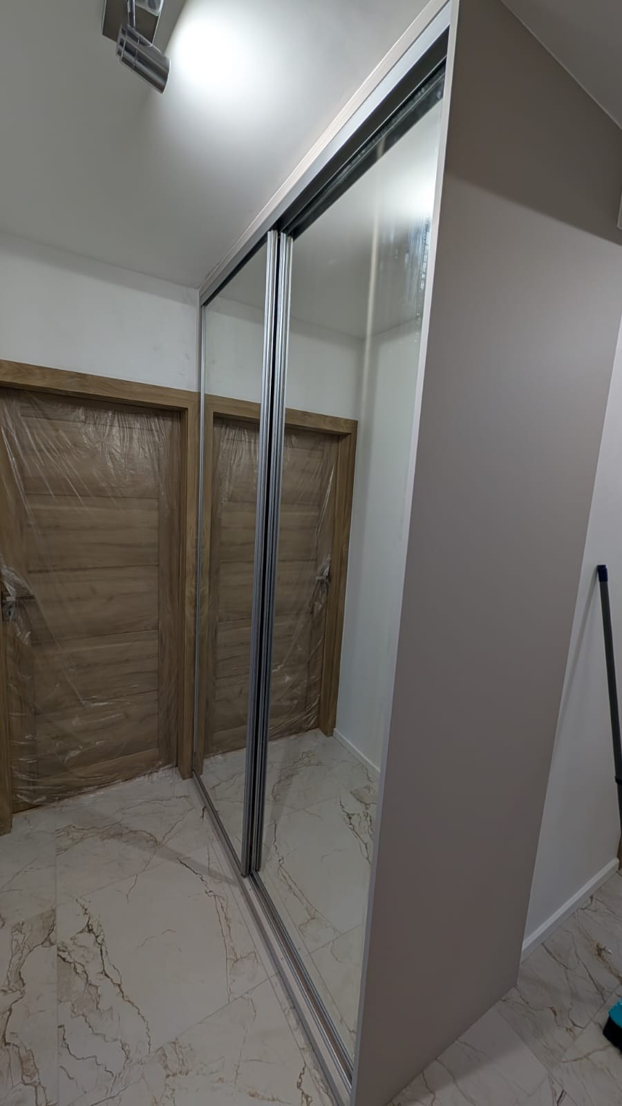 Jasna szafa wnękowa z lustrzanymi drzwiami przesuwnymi w Opalenicy. Widoczne drzwi zabezpieczone folią ochronną, marmurowa podłoga i fragment ściany.