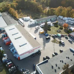 Widok z lotu ptaka na hurtownię budowlaną MAK-LER z zaparkowanymi samochodami i składowanymi materiałami budowlanymi, otoczoną jesiennymi drzewami.