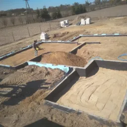 Wykop pod fundamenty domu jednorodzinnego, zabezpieczony izolacją termiczną (styropian) i przeciwwilgociową, z widocznym pracownikiem zagęszczającym podłoże ubijarką wibracyjną.