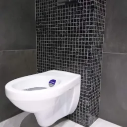 Nowoczesna toaleta podwieszana marki Cersanit na tle ściany wyłożonej czarną mozaiką z błyszczącymi elementami, z chromowanym przyciskiem spłukującym.