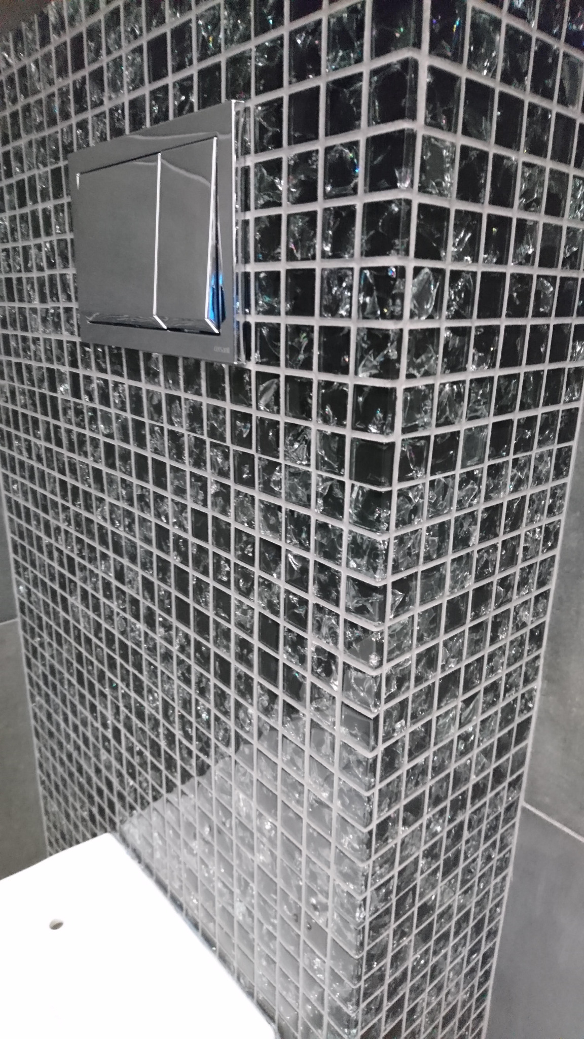 Ściana obłożona czarną mozaiką szklaną z chromowanym przyciskiem spłuczki, widok z dołu, fragment białej umywalki.