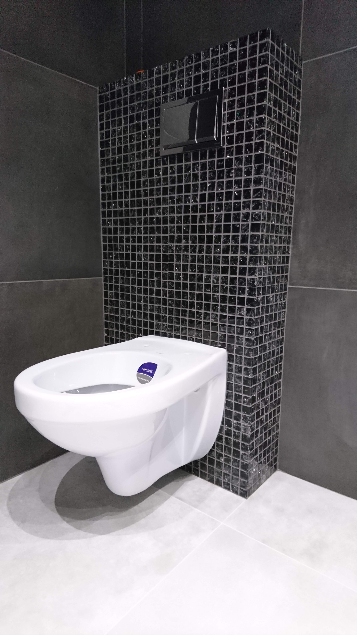 Nowoczesna toaleta podwieszana marki Cersanit na tle ściany wyłożonej czarną mozaiką z błyszczącymi elementami, z chromowanym przyciskiem spłukującym.