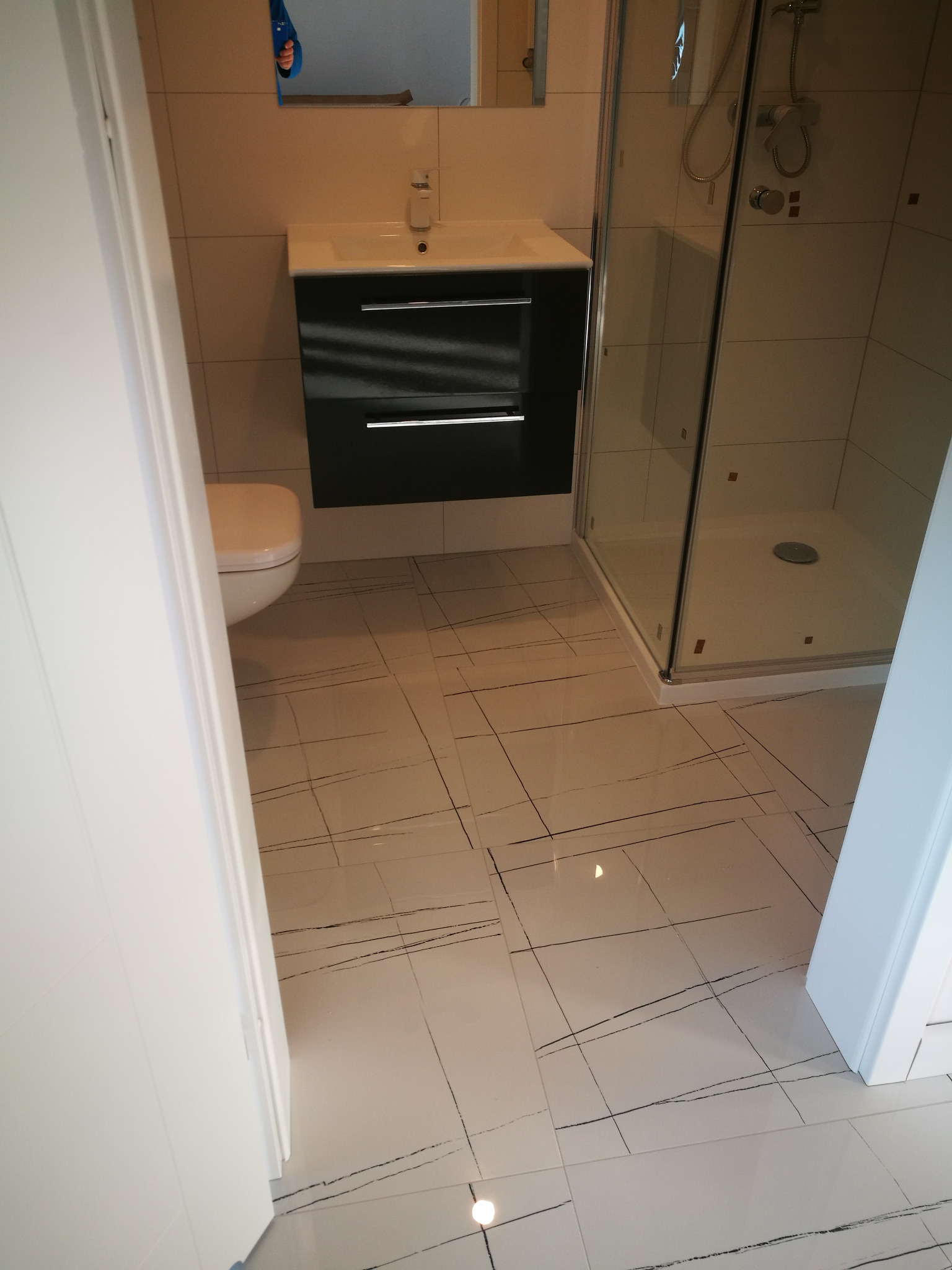 Nowoczesna łazienka z kabiną prysznicową, umywalką z szafką w kolorze czarnym i toaletą, wykończona białymi płytkami podłogowymi z czarnymi żyłkami.