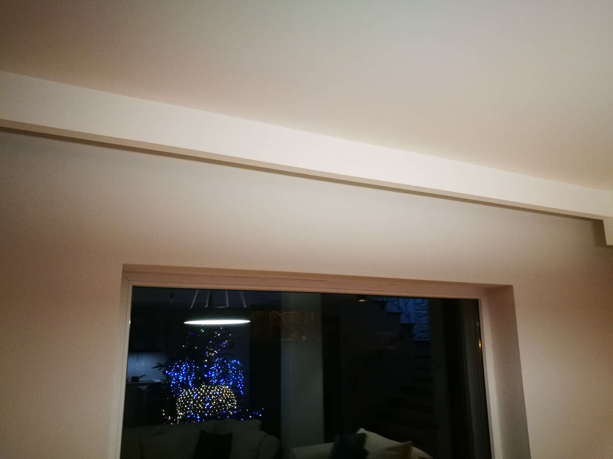 Ściana pomalowana na jasny odcień szarości z białym sufitem i listwą przysufitową; widok przez okno na choinkę w tle.