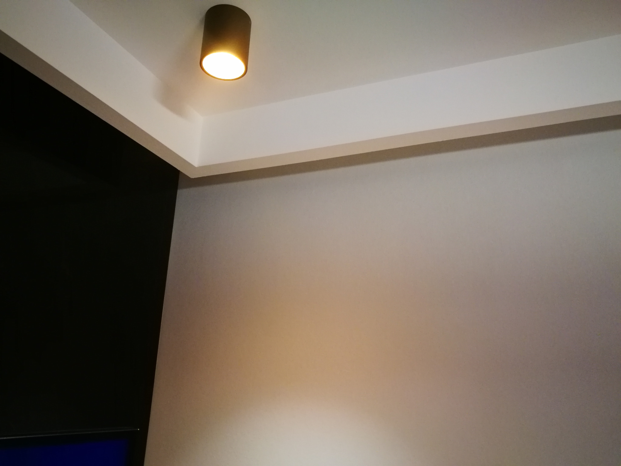 Zamontowana okrągła lampa sufitowa wpuszczana w narożniku pomieszczenia z szarą ścianą i czarnym elementem wykończenia.