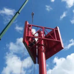 Montaż konstrukcji reklamowej na słupie z użyciem dźwigu, pracownik w kasku zabezpieczony linami na wysokości, błękitne niebo z chmurami w tle.