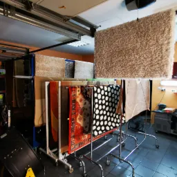 Różnorodne dywany, w tym wzorzyste i puszyste, suszone na metalowych stojakach w pomieszczeniu z pomarańczowymi ścianami i szarą podłogą, widoczny fragment sufitu garażowego.