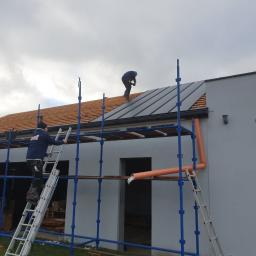 POKRYCIA DACHOWE - BARTŁOMIEJ LEŚNIAK - Dwóch dekarzy pracuje na rusztowaniu przy montażu blachy na dachu domu jednorodzinnego, widoczna więźba dachowa i częściowo ułożona blacha w kolorze grafitowym.