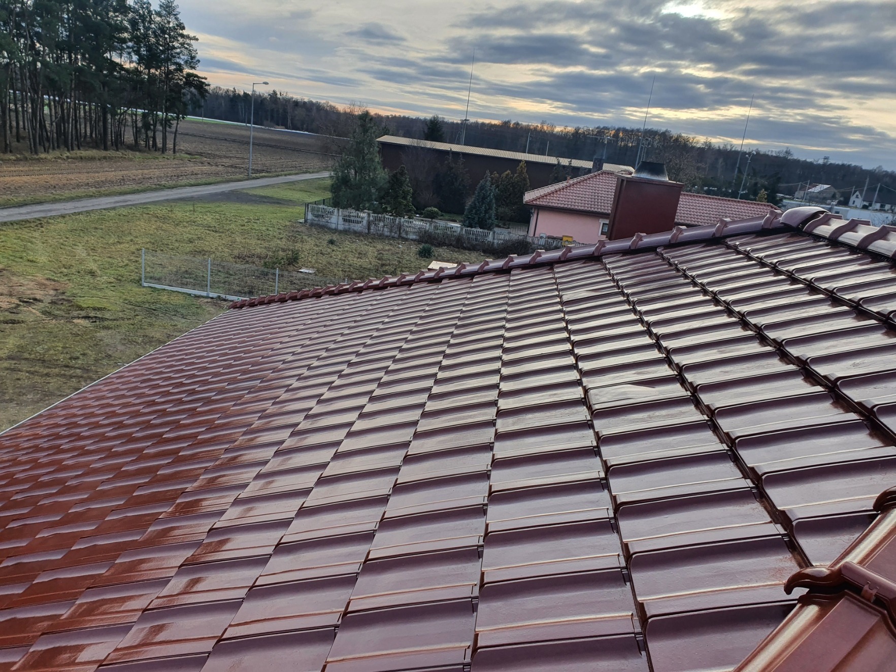 Nowy, błyszczący dach z dachówki ceramicznej w kolorze brązowym, widok z góry na tle krajobrazu wiejskiego z drzewami i budynkami w oddali.